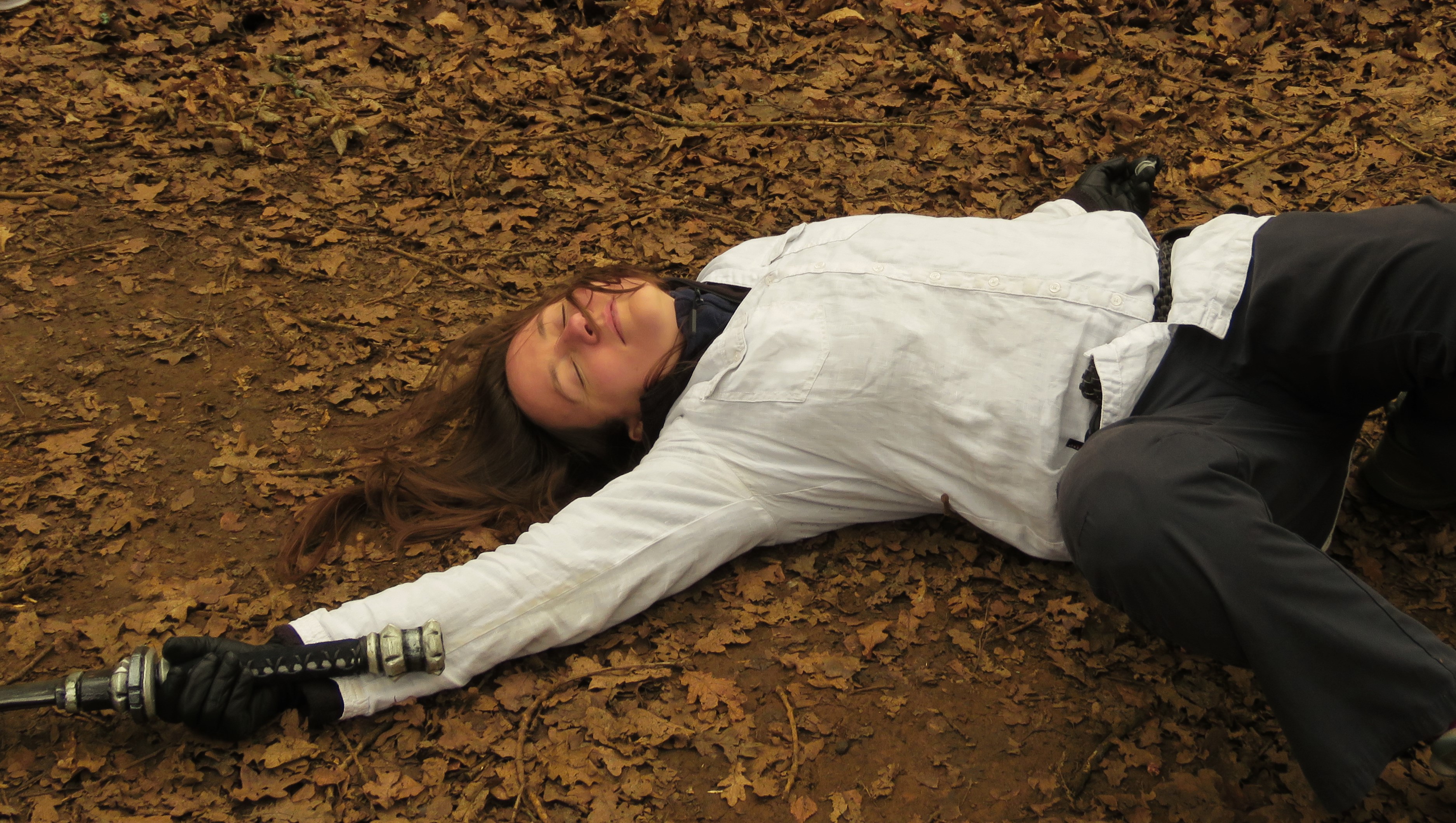  A player in a white shirt collapsed on a path strewn with brown leaves, weapon hand outstretched.
